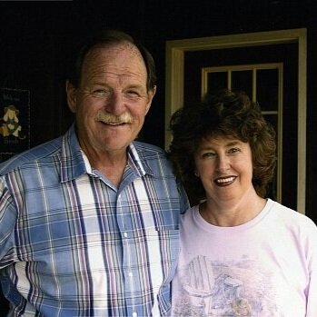 Smiling elderly couple standing together outdoors, wearing casual clothing; the man has a plaid shirt and the woman has a light-colored shirt.