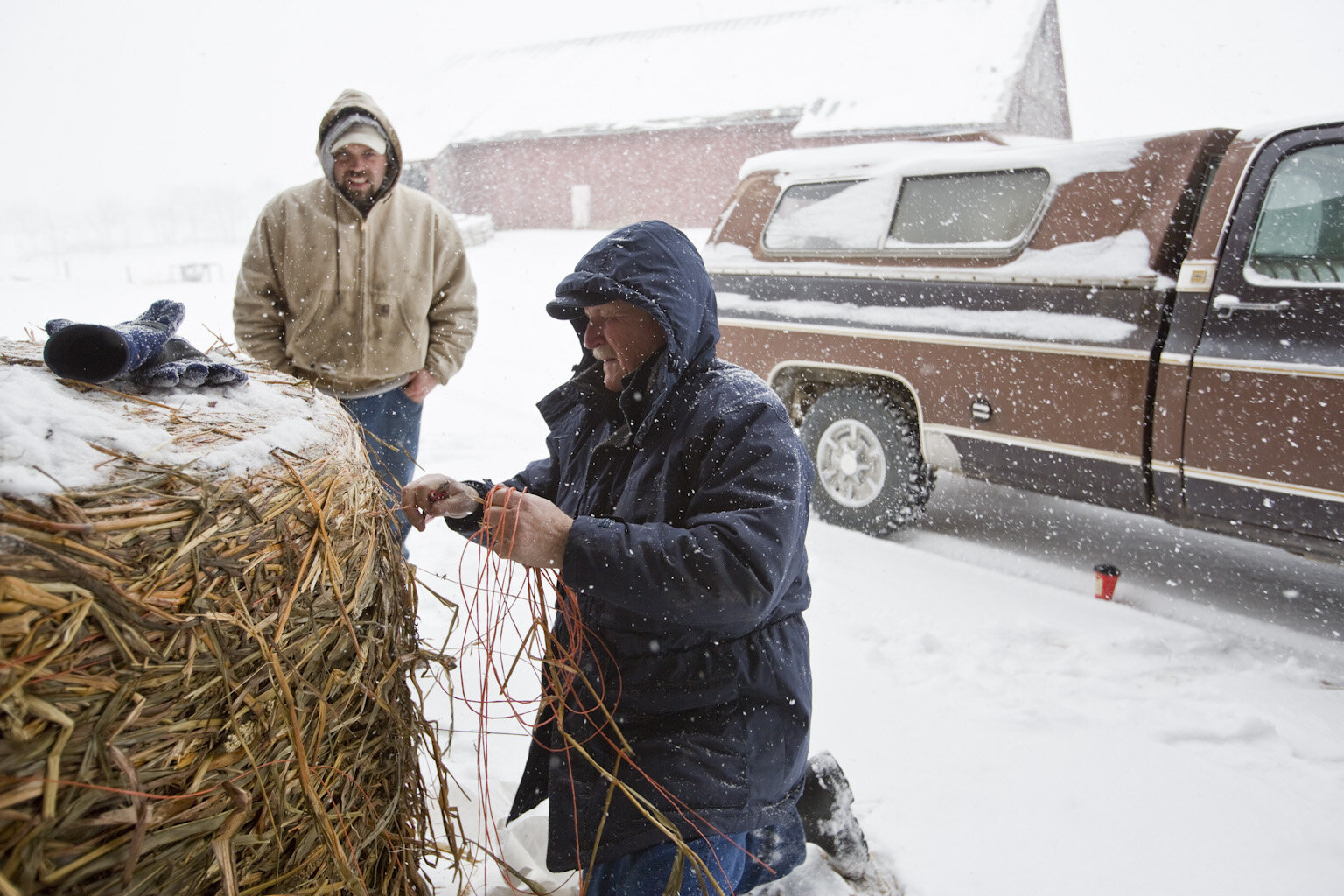 Two men working outdoors in heavy snow, one kneeling by a straw bale tying wires, while the other stands nearby observing. A brown pickup truck and a barn are visible in the background, creating a rural winter farming scene.