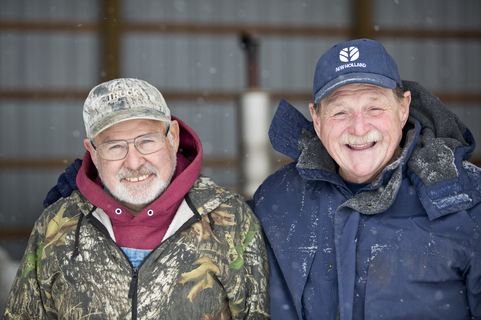 Two smiling older men standing together outdoors in winter clothing, with snow gently falling around them. One man is wearing a camouflage jacket and a cap, while the other is dressed in a dark blue coat and a matching cap. The background features a barn-like structure, indicating a rural setting.