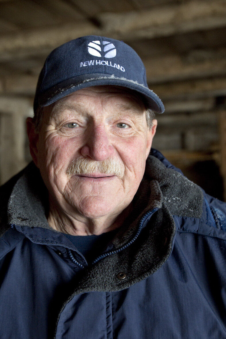 Portrait of an elderly farmer wearing a blue New Holland cap and a dark jacket, smiling warmly in a rustic barn setting. The image captures the essence of rural life and agricultural heritage.