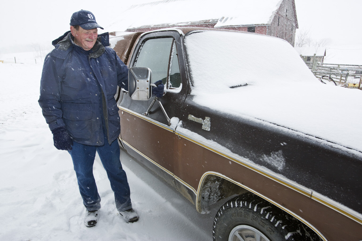 Man standing next to a snow-covered brown pickup truck in a winter landscape, wearing a blue jacket and hat, with snow falling in the background. A barn is visible in the distance.