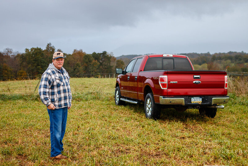 A man in a plaid shirt and cap stands in a grassy field next to a red Ford truck, surrounded by trees under a cloudy sky, showcasing rural life and farming culture.