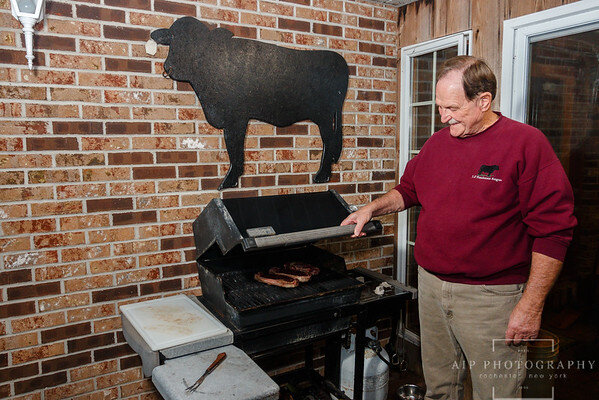 Man grilling steaks on a barbecue grill in a backyard with a cow silhouette decoration on the wall, showcasing outdoor cooking and grilling techniques.