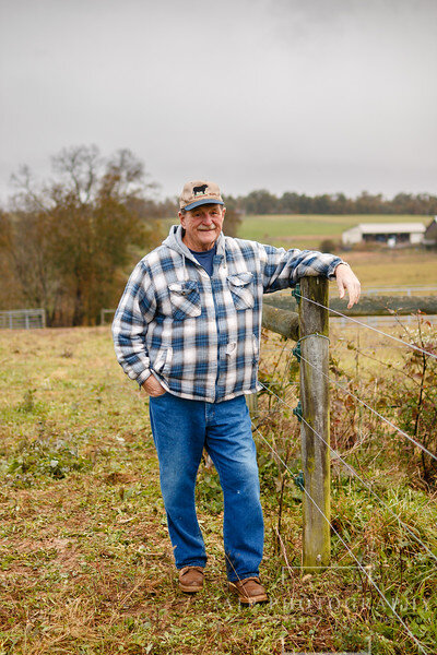Smiling farmer in a plaid shirt and baseball cap stands by a wooden fence in a rural landscape, with fields and a barn in the background, showcasing agricultural life.