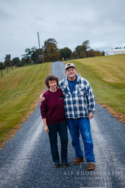 A couple standing together on a rural road, surrounded by green fields under a cloudy sky, showcasing a warm and friendly moment in a serene countryside setting.