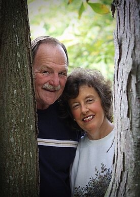 A smiling older couple posing together between two trees, surrounded by greenery, showcasing a joyful moment in nature.