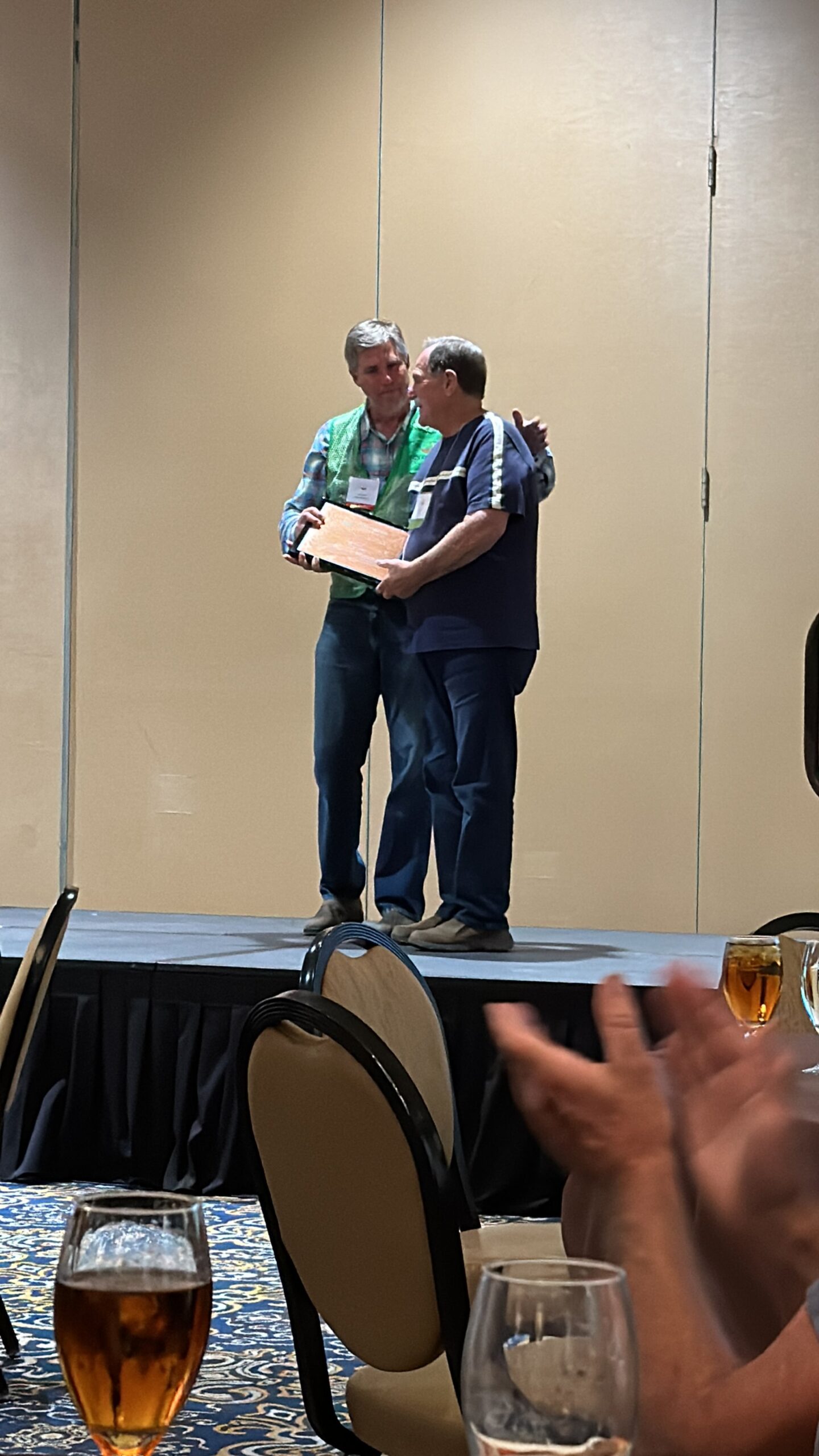 Two men on stage at an event, one presenting an award or plaque to the other. The audience is visible in the foreground, with drinks on the table. The setting features a neutral backdrop and a formal atmosphere, indicating a celebration or recognition ceremony.
