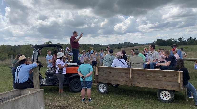 Group of people gathered outdoors for an informational session, with a speaker on a utility vehicle addressing the audience. The setting features a farm landscape under cloudy skies, showcasing a diverse group engaged in discussion about agriculture or sustainability.