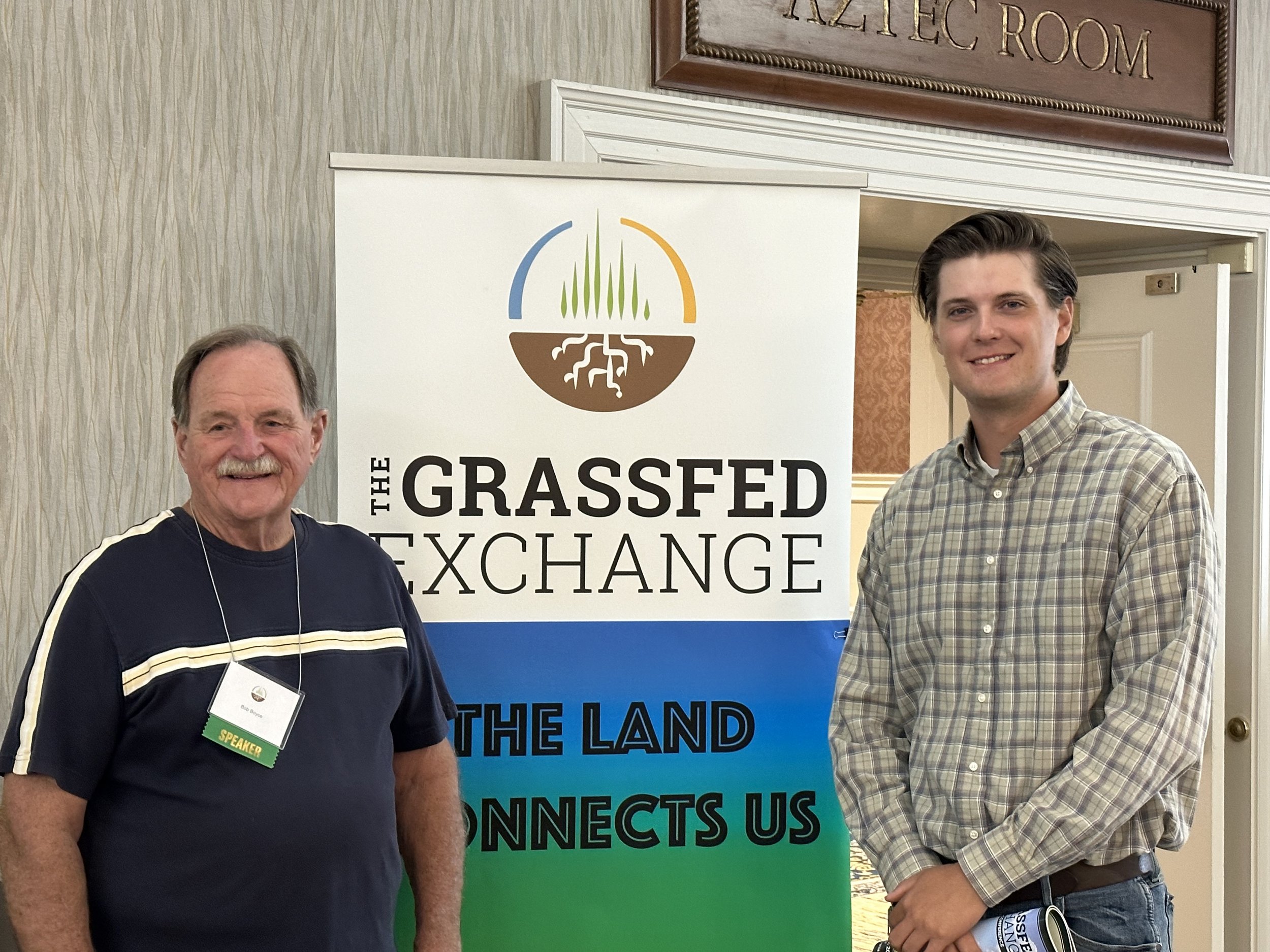 Two men standing in front of a banner for The Grassfed Exchange, which features the slogan "The Land Connects Us." The man on the left is wearing a navy shirt with white stripes and a name tag indicating he is a speaker. The man on the right is dressed in a plaid shirt. The setting appears to be a conference or event space.